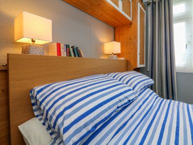 A bedroom with a bed with blue and white striped bedding two lamps on a wooden headboard and a window with blue curtains at 4 The Park in Kilkhampton