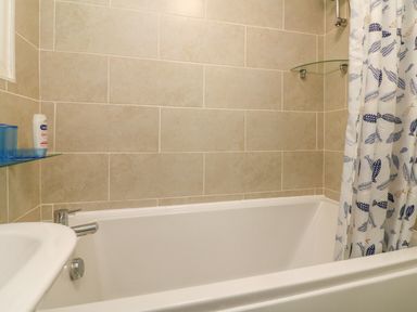 A bathroom with a white bathtub beige tiled walls a shower curtain with fish patterns and a small glass shelf holding toiletries at 4 The Park in Kilkhampton