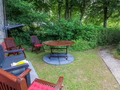 An outdoor garden area with wooden chairs and a round table on grass next to a paved pathway at 4 The Park in Kilkhampton