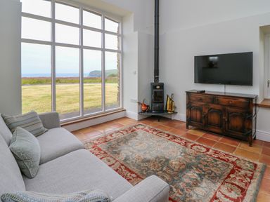 A living room with a large window, a gray sofa, a patterned rug, a wood stove, and a television on a wooden cabinet at West Dairy near Newport Pembrokeshire