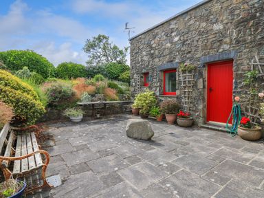 A stone patio with a bench and potted plants outside a stone building with red door and windows at West Dairy near Newport Pembrokeshire