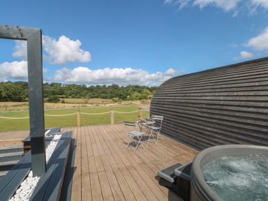A wooden deck with a hot tub small table and chairs next to a curved wooden building and a grassy field at Paradise in Kenstone near Hodnet