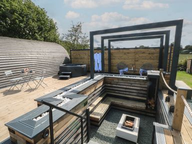 An outdoor wooden deck with a sunken seating area around a fire pit a hot tub small dining table two chairs and a privacy screen at Paradise Kenstone near Hodnet