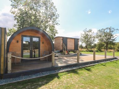 An outdoor wooden pod with glass doors and chairs on a deck next to a small wooden cabin on stilts at Hawkstone Kenstone near Hodnet