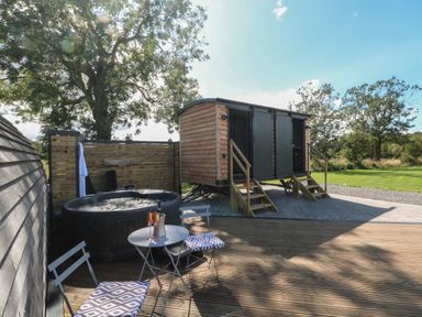 An outdoor wooden deck with a hot tub two chairs and a small table with glasses next to a wooden cabin at Hawkstone Kenstone near Hodnet