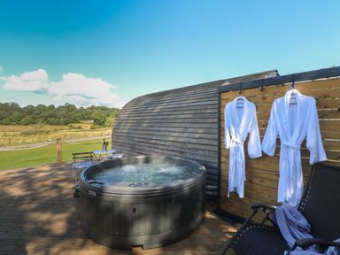 An outdoor wooden deck with a round hot tub two white bathrobes hanging on a wooden wall and outdoor chairs at Hawkstone in Kenstone near Hodnet