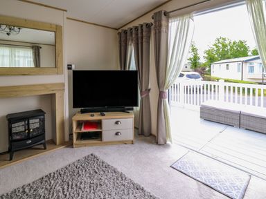 A living room with a television on a wooden stand next to curtains and a door leading to a deck with seating at Osprey 6 in Kiln Park Tenby