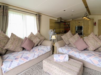 A living room with two sofas, cushions, a small table lamp, a fan, an ottoman and a kitchen area in the background at Osprey 6 Kiln Park Tenby