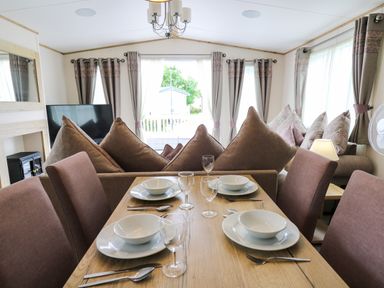 A dining table set with plates bowls glasses and cutlery next to a living area with sofas and curtains at Osprey 6 Kiln Park Tenby