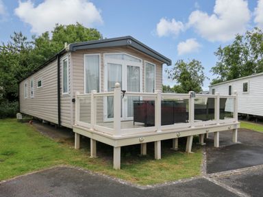 A beige mobile home with a glass-railed deck and outdoor furniture on grass at Osprey 7 in Tenby