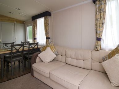A living room with a beige sofa with cushions and a dining table with six chairs by windows with yellow floral curtains at Osprey 7 in Tenby