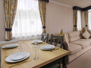 A dining table set with plates and glasses next to a beige sofa with cushions in a room with patterned curtains at Osprey 7 in Tenby