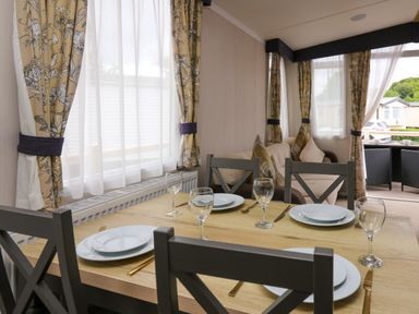 A dining table set with plates and glasses next to a sofa and windows with patterned curtains at Osprey 7 in Tenby