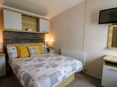 A bedroom with a double bed with patterned bedding and yellow pillows two bedside tables with lamps a wall-mounted cabinet and a television at Osprey 7 in Tenby