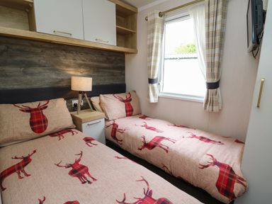 A bedroom with two single beds with red deer-patterned bedding a bedside table with a lamp a window with plaid curtains and wall cabinets at Osprey 7 in Tenby