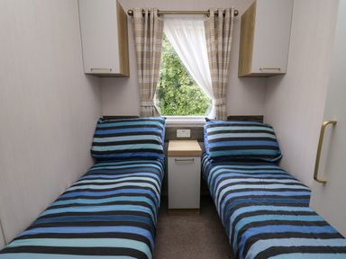 A small bedroom with two single beds with striped blue bedding a window with curtains and a small cabinet between the beds at Osprey 7 in Tenby