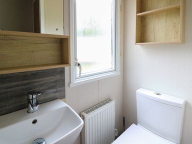 A bathroom with a white sink, toilet, radiator, a window, and wooden shelves at Osprey 7 in Tenby
