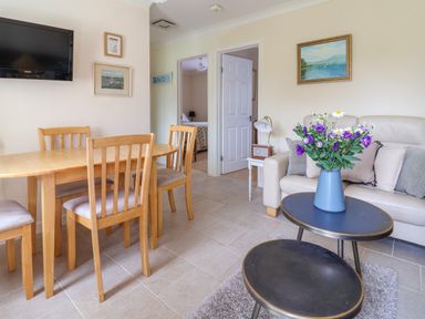 A dining table with four wooden chairs a wall mounted TV a sofa with cushions and a coffee table with a vase of purple flowers at Seaweed in Weston near Sidmouth