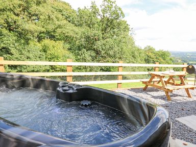 An outdoor hot tub with water bubbling next to a wooden picnic table and a barbecue grill with trees and hills in the background at Shepherd's Cwtch in Cilycwm near Llandovery