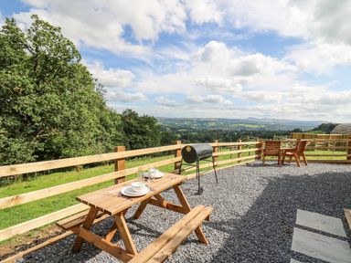 An outdoor seating area with a wooden picnic table set with dishes a barbecue grill and wooden chairs overlooking a green landscape with trees and hills at Shepherd's Cwtch in Cilycwm near Llandovery