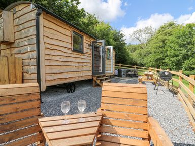 An outdoor area with wooden chairs and table with glasses a wooden picnic table a barbecue grill and a wooden cabin at Shepherd's Cwtch in Cilycwm near Llandovery