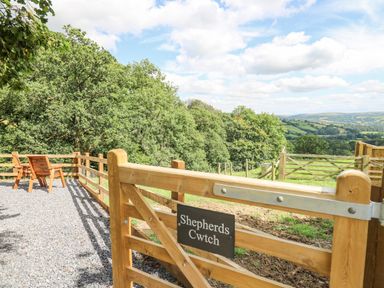 A wooden gate with a sign Shepherds Cwtch overlooking a green hilly landscape with wooden chairs on a gravel area at Shepherds Cwtch in Cilycwm near Llandovery