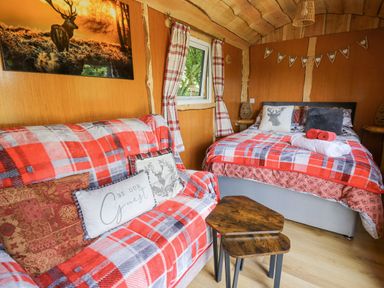 A small room with a bed, a sofa covered in red and gray plaid blankets, a small wooden table, and a window with plaid curtains at Shepherd's Cwtch in Cilycwm near Llandovery