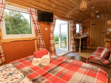 An interior room with a bed covered with a plaid blanket and rolled towels a mounted television a door leading outside a small table with stools and a couch with plaid covers at Shepherd's Cwtch in Cilycwm near Llandovery