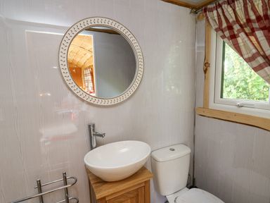 A bathroom with a round mirror above a sink a toilet and a window with checked curtains at Shepherd's Cwtch in Cilycwm near Llandovery