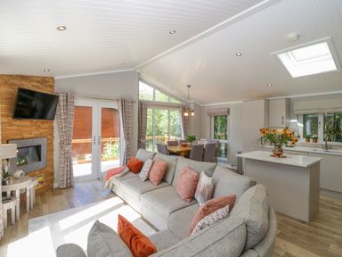 An open living area with a grey sofa and patterned cushions a dining table with chairs and a kitchen island with flowers at Waterside Lodge 55A Bowness White Cross Bay near Windermere