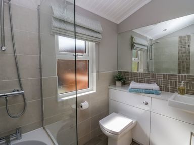 A bathroom with a shower over a bathtub a window with a blind a toilet and a sink with towels and a plant on the counter at Waterside Lodge 55A Bowness White Cross Bay near Windermere