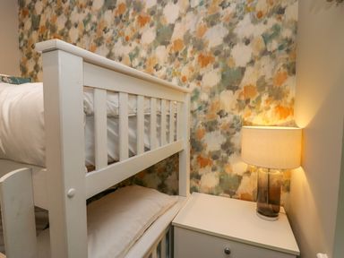 A white bunk bed next to a white nightstand with a lamp and a multicolored abstract wallpaper in a bedroom at Waterside Lodge 55A Bowness White Cross Bay near Windermere
