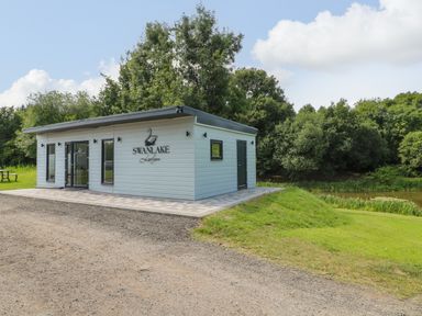 A single story white building with swanlake lodges sign near a pond and trees at Peacock Lodge in Swannington near Coalville