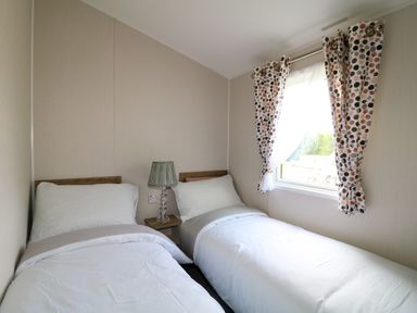 A bedroom with two single beds a nightstand with a lamp and a window with patterned curtains at Peacock Lodge in Swannington near Coalville