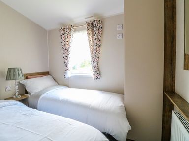 A bedroom with two single beds white bedding a bedside table with a lamp and a window with patterned curtains at Peacock Lodge in Swannington near Coalville