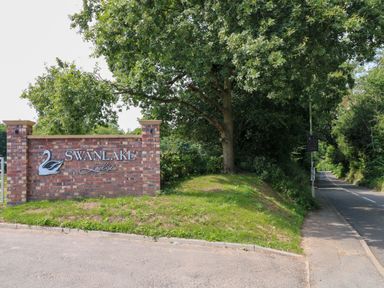 A brick sign for Swanlake Lodges next to a road with a large tree and greenery at Peacock Lodge in Swannington near Coalville
