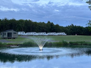 A pond with a water fountain in front of several mobile homes and trees at Peacock Lodge in Swannington near Coalville