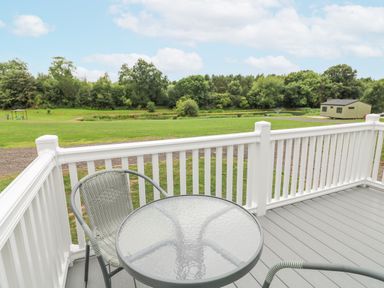 A deck with a glass table and two chairs overlooking a grassy area and trees at Peacock Lodge in Swannington near Coalville