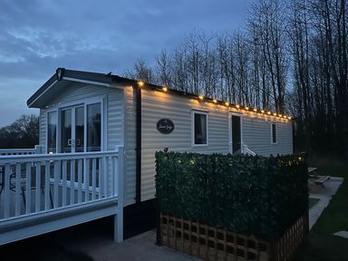 A mobile home with outdoor string lights and a white railing surrounded by trees at Peacock Lodge in Swannington near Coalville
