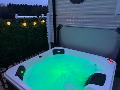 An outdoor hot tub with illuminated water and headrests near a white wall and a fence with lights at Peacock Lodge in Swannington near Coalville