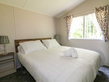 A bedroom with a double bed white bedding two bedside tables with lamps and a window with patterned curtains at Stork Lodge in Swannington near Coalville