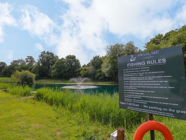 A fishing lake with a fountain surrounded by trees and a fishing rules sign at Swanlake Lodges