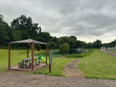 A wooden shelter with picnic table and benches next to playground equipment by a pond with a fountain near trees at Stork Lodge in Swannington near Coalville