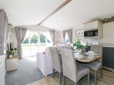 An open living and dining area with a dining table set with dishes and a vase of flowers a sofa a TV and glass doors leading outside at Stork Lodge in Swannington near Coalville