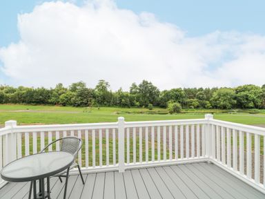 A deck with a round glass table and one chair overlooking a grassy field and trees at Stork Lodge in Swannington near Coalville