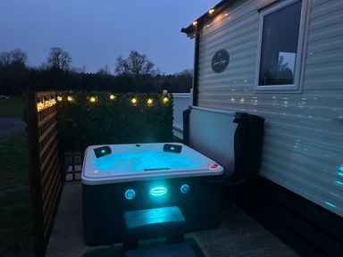 An outdoor hot tub next to a mobile home with string lights and greenery at Stork Lodge in Swannington near Coalville