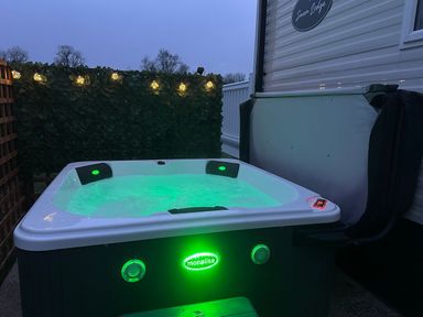 An outdoor hot tub with green lights next to a mobile home with a privacy screen and string lights at Swan Lodge in Swannington near Coalville