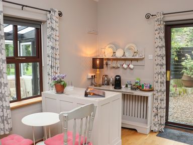A small dining area with a white table and chairs with pink cushions near windows and a shelf with plates and cups at Pink Granite Garden of Ireland near Tinahely County Wicklow