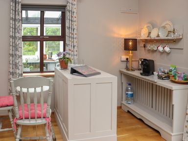 A kitchen area with a white table, chairs with pink cushions, window with patterned curtains, coffee machine, lamp, and shelves with plates and cups at Pink Granite Garden of Ireland near Tinahely County Wicklow