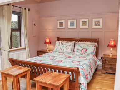 A bedroom with a wooden bed and floral bedding two wooden stools window with curtains and bedside tables with lamps at Pink Granite Garden of Ireland near Tinahely County Wicklow